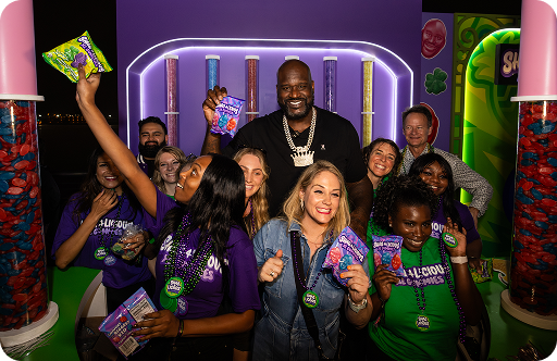 Group of people celebrating with candy at a colorful event, holding candy bags and smiling in front of a purple backdrop.