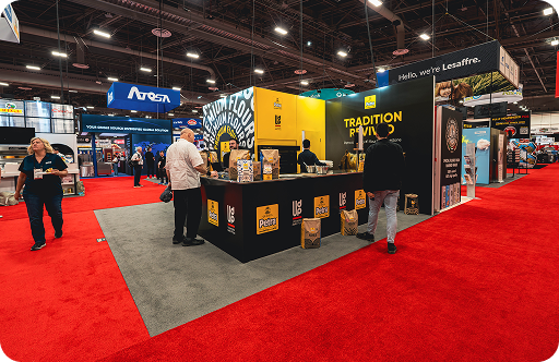 Trade show booth with black and yellow branding for Petru flour featuring product displays and attendees on a red carpet floor.