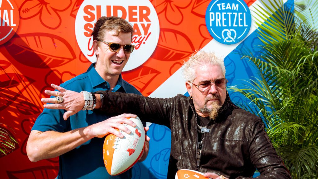 Two men hold footballs and pose in front of a colorful King’s Hawaiian backdrop featuring “Team Original” and “Team Pretzel” signage.