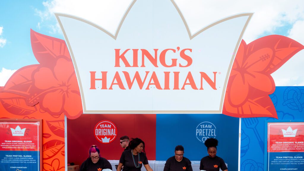 King’s Hawaiian booth with staff serving food under a large crown-shaped sign featuring “Team Original” and “Team Pretzel” sections.